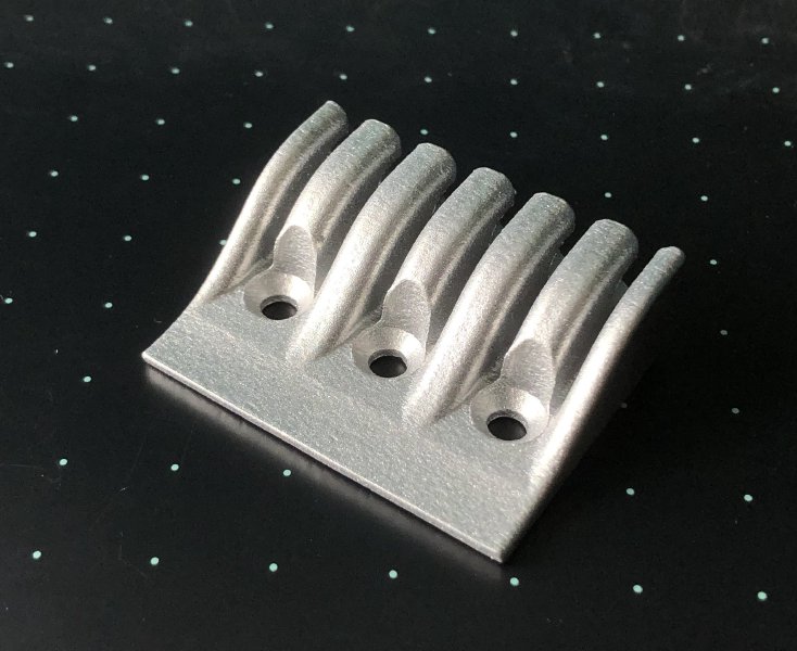 A photo of a 3D-printed aluminum part that will form the headstock of my new guitra. It has a small rectangular base about 42mm across, with holes for 3 screws, and then seven fins that raise up to trap the string ends.