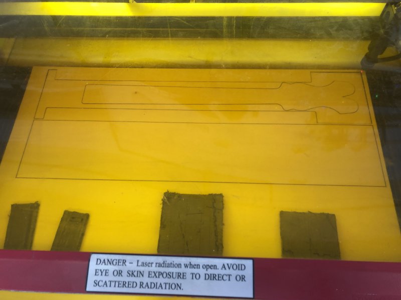 A sheet of plywood rests in the laser cutter, and you can see the outline of a long rectangle cut (the base) and another I beam shaped piece with a guitar neck profile cut in it.