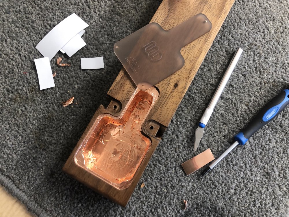 The rear body of the guitar on a carpet mat on a workbench, showing the copper covered inside of the electronics cavity. To the side you can see a scalpel and bits of tape, and on the body to one side you can see the translucent orange acrylic cover for the cavity that will let the shiny copper inside be visible once the cover is on.