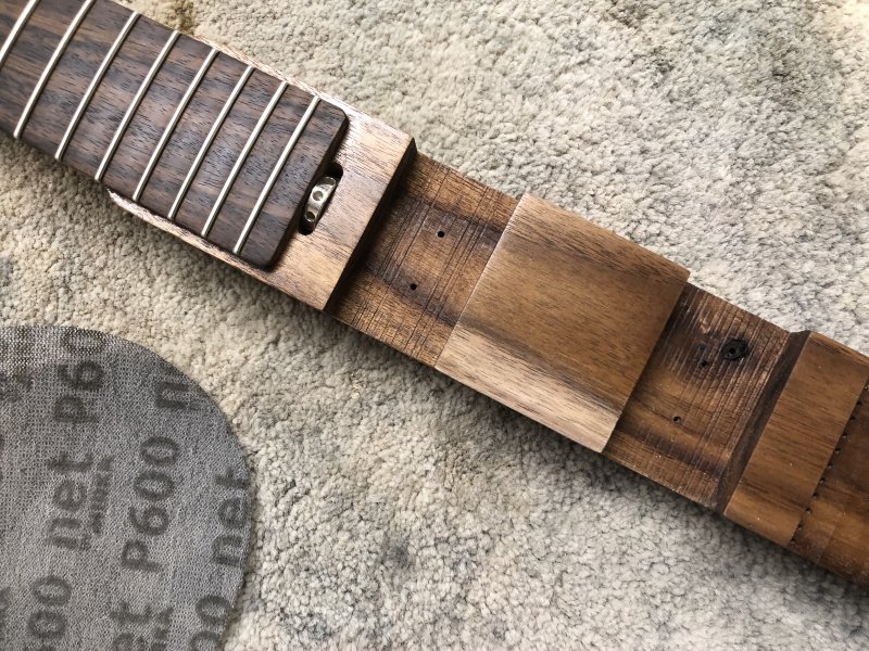 A close up on the mid point of Älgen guitar's body, which is mostly a dark natural wood, except for one patch which is lighter due to having been sanded back. Beside the body is a 600 grit sanding disk.