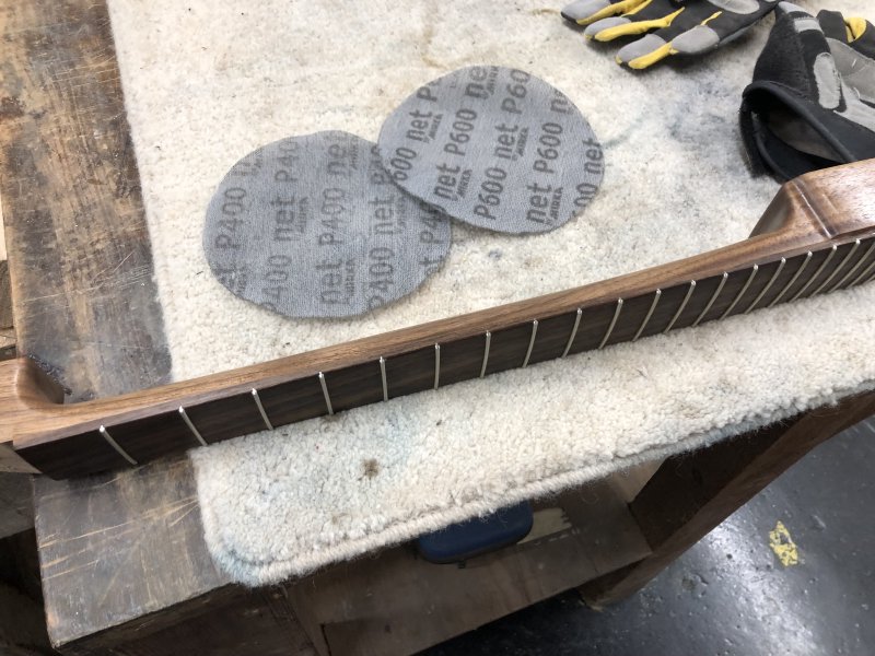 The side of the neck of the guitar is seen on the workbench, with two sanding disks beside it.