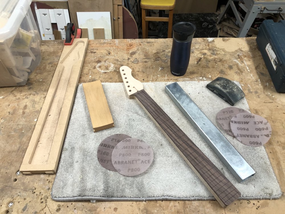 A work in progress guitar neck sits on the workbench, surrounded by a metal beam about a foot long that is used for testing straightness, sanding disks and sanding blocks.