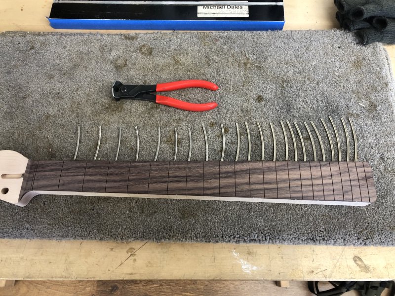 The neck on the workbench with a strip of fretwire placed next to each empty fret-slot. Besides them is the cutter I used to snip the fretwire off the reel. 