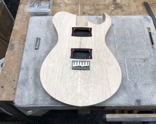 A guitar body sat on the workshop, with a couple of parts laid on top of it to give the impression of how it might look when finished. The top is a nice birds-eye maple, and on one side it is nicely rounded, and on the other side it has a pointy horn to it.