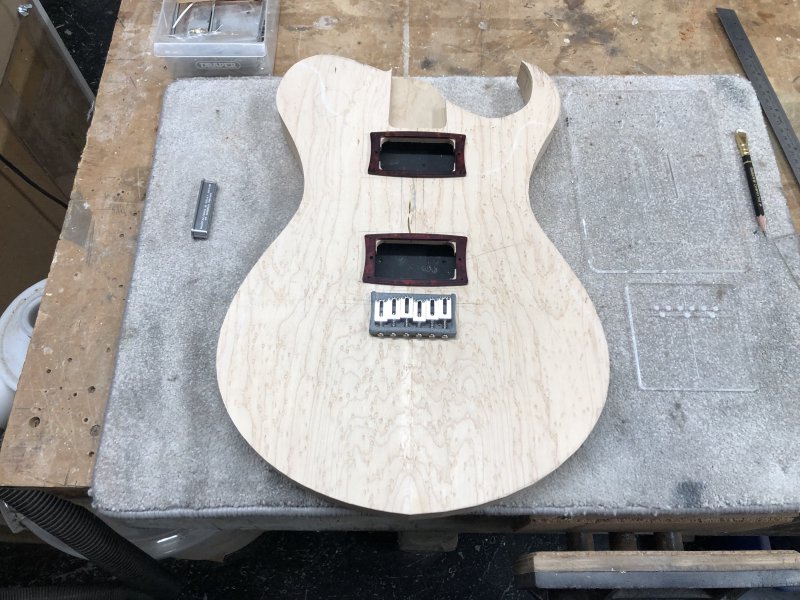 A guitar body sat on the workshop, with a couple of parts laid on top of it to give the impression of how it might look when finished. The top is a nice birds-eye maple, and on one side it is nicely rounded, and on the other side it has a pointy horn to it.