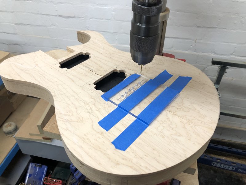 A guitar body mounted on a drill press with a laser-cut acrylic guide taped to the front to let me line up all the holes I need to drill.