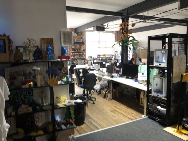 A wide shot of the main maker room at DoES Liverpool, with a lots of things people have made there on shelves to the left, a rack containing two 3D printers to the right, and lots of benches with monitors on in between.