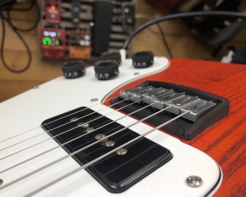 The 3D printed bridge mounted on the guitar with strings running over it. In the background you can see the floor and my pedal board, telling you that this is now being used!