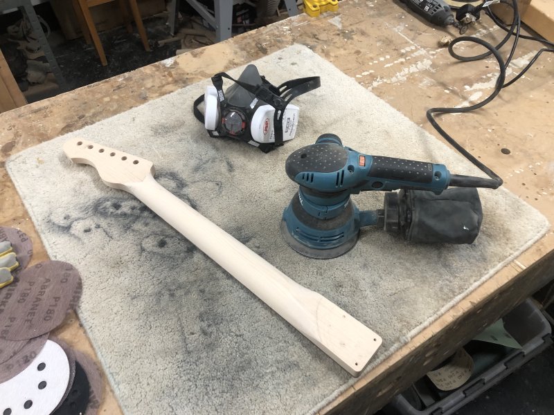 The neck now sits on a carpet map on a workbench, looking deceptively close to being complete thanks to the poor photo. Beside the neck sits an makita orbital sander, a face mask, and some sanding disks.