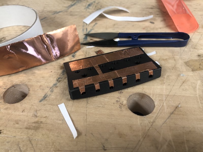 The 3D-printed bridge part is on the workbench with bits of copper tape all over the under-side, with six fingers of tape running up the back edge to where the saddles attach. Surrounding the bridge is a bunch of tape and some cutters.