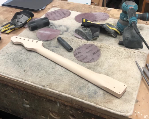 An unfinished guitar neck sits fretboard down on a workbench, surrounded with sanding disks.