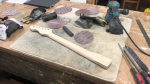 An unfinished guitar neck sits fretboard down on a workbench, surrounded with sanding disks.