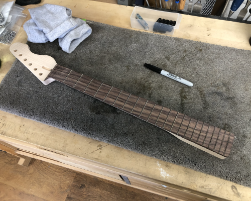 A work in progress guitar neck, shaped and fretted, sat on a workbench. Beside the neck is a black sharpie pen, and closer inspection reveals all the frets have been drawn on with the sharpie ready for fret shaping work.
