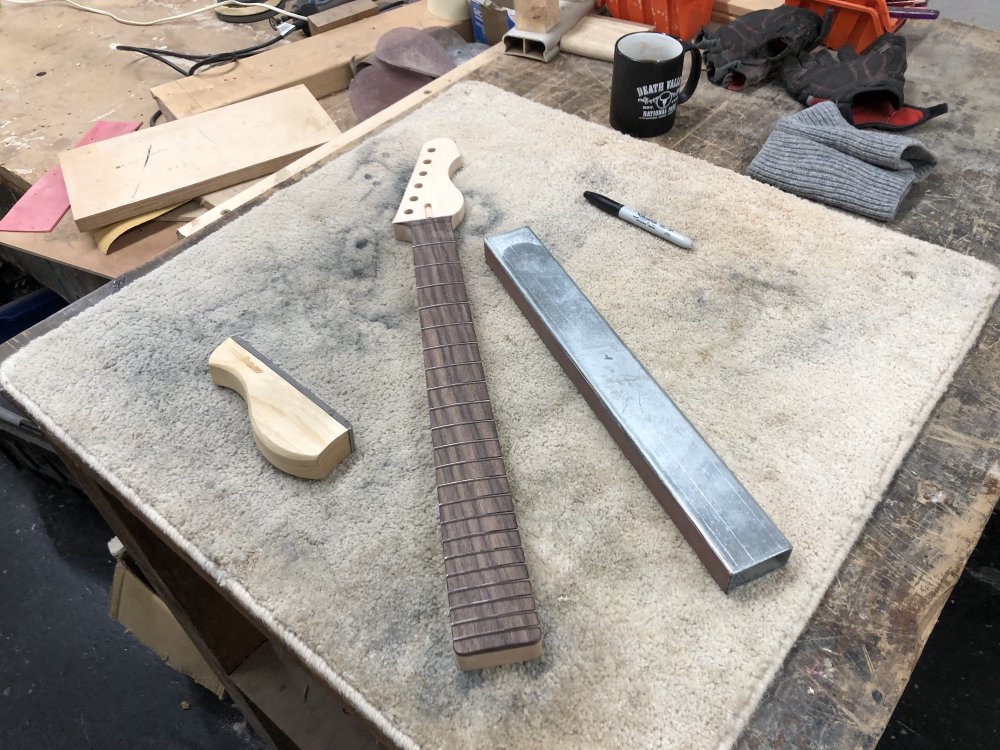 A unfinished guitar neck sits on a carpet mat on a workbench, around which sits a small hand file, a sanding bar (fancy name for a long straight bit of metal with sandpaper on one side), and a black sharpie pen.