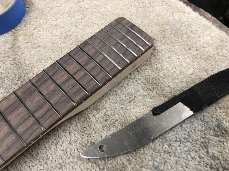 A close up of the guitar-end of the neck, showing the frets with black sharpie drawn all over them, and next to that sits a metal tool that is the fret-crowning file.