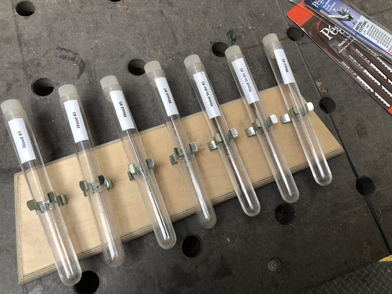 A series of plastic test-tubes attached to a plank of wood, each one has a label on it for the type of scroll saw blade that is held within.
