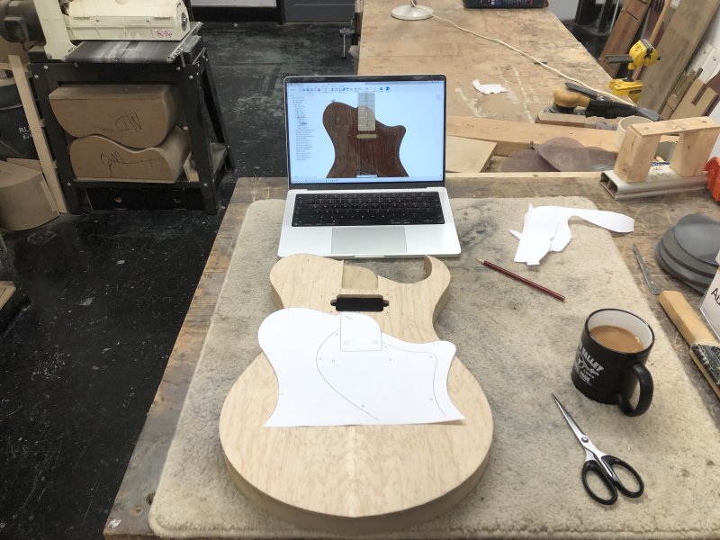 The body of an existing guitar that's being built sits on the desk, over which is a paper cutout of the new design laid over top.