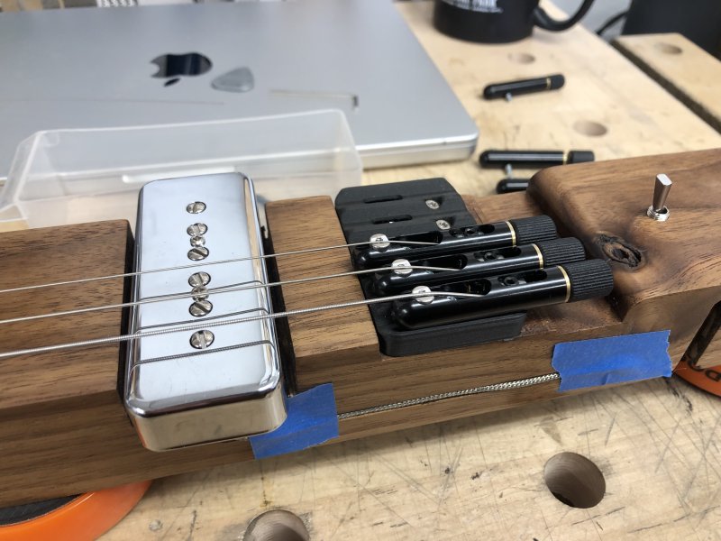 The bridge of the guitar is shown, with three of the tuning units installed and strung up, with the rest seen off to one side. The tuning units are black tubes with hemispherical ends, vaguely looking like a submarine.