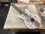 An unusual looking guitar sat on a workbench. The guitar consists of a wooden central core that consists of the neck extends that profile through the body, and then the sides of the body are made from a 3D printed lattice. The guitar has no headstock, instead having tuners at the bridge.