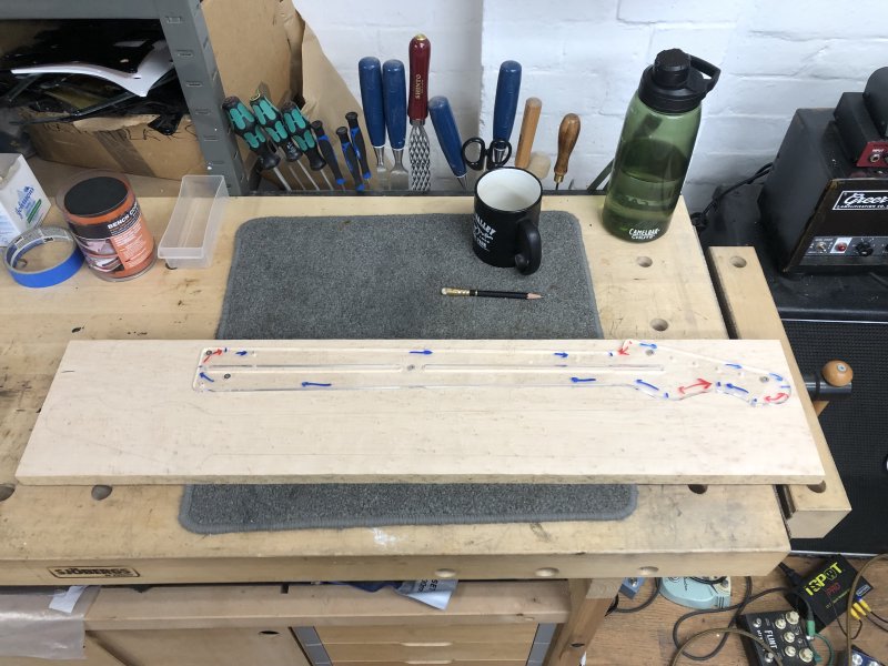 A plank of birds-eye maple sits on the workbench, upon which sits one neck template, and you can see a pencil marking where a second neck could go.