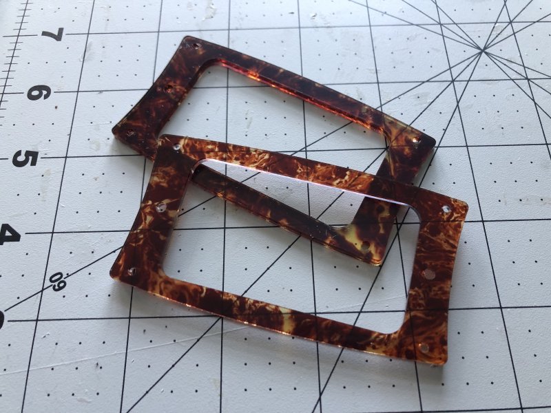 A pair of pickup rings (aka, slightly rounded rectangle frames with some screw holes in) sit on a craft mat. They are made from a translucent reddish turtleshell effect material.