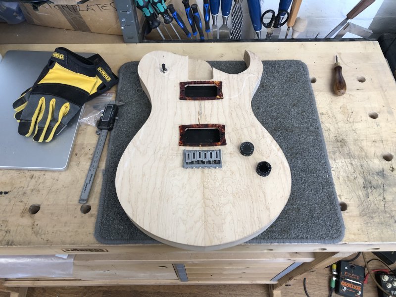 A work in progress guitar body sits on the workbench, and placed on top of it are the bridge, volume/tone controls, pickup rings, etc.