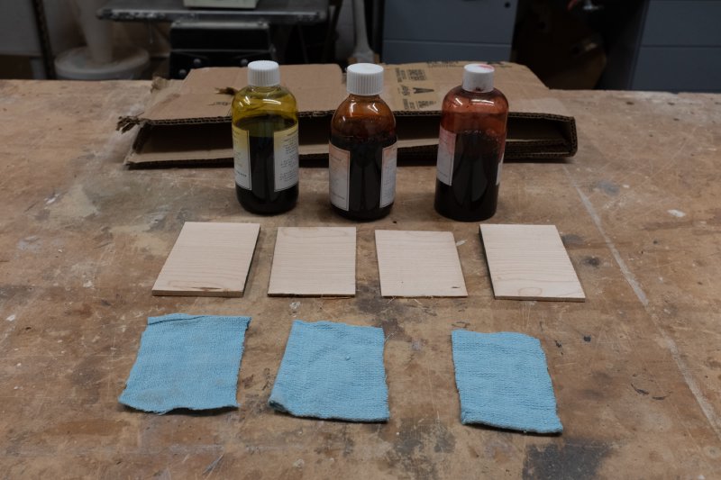 Three bottles of stain sit behind four postcard sized bits of wood, and in front of those are three bits of cloth ready to use as applicators.
