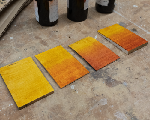A set of four postcard-sized rectangles of maple on a workbench, each with a different stain blend being tested: first is just straight yello, the second is orange over yellow, the third is red over yellow, and then the last is yellow, orange, and red.