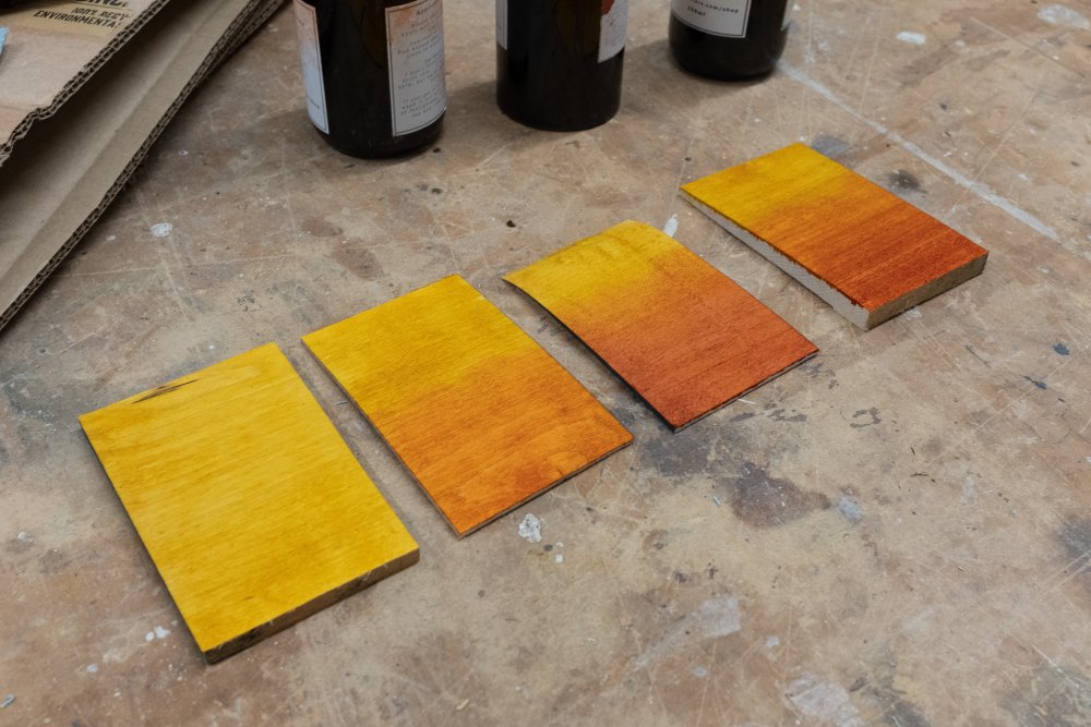 A set of four postcard-sized rectangles of maple on a workbench, each with a different stain blend being tested: first is just straight yello, the second is orange over yellow, the third is red over yellow, and then the last is yellow, orange, and red.