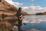 Me standing holding a guitar in the Snowdia landscape, with a hill to my left and a lake to my right.