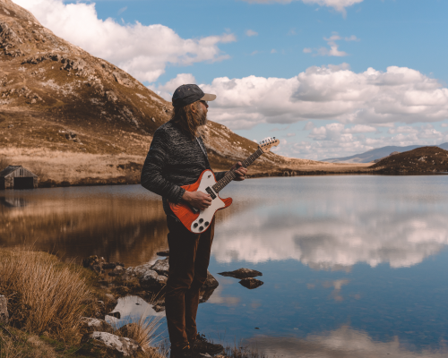 Me standing holding a guitar in the Snowdia landscape, with a hill to my left and a lake to my right.