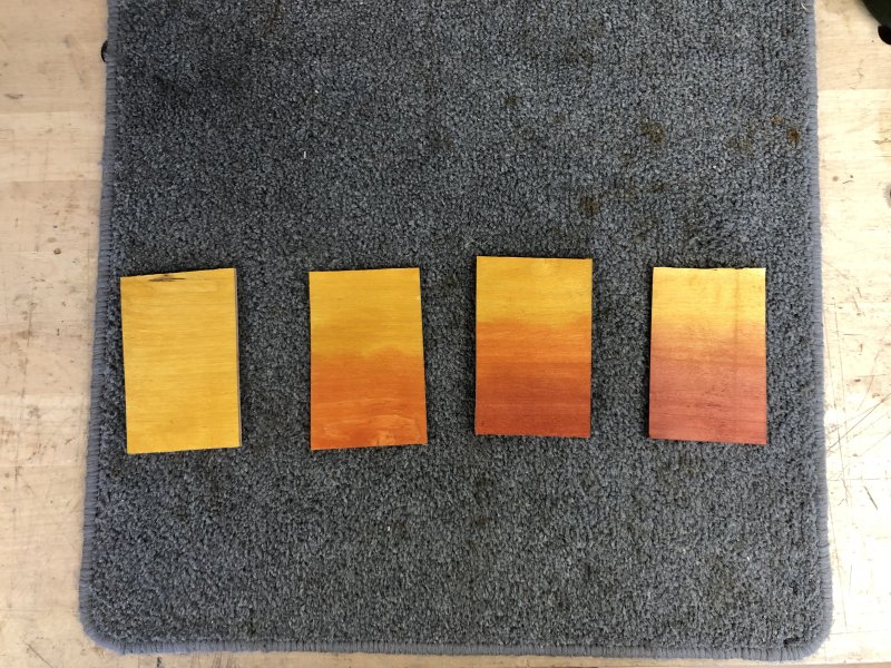 Four postcard sized bits of wood sit on the workbench, each with a different set of strains: all yellow, orange faded to yellow, red faded to orange to yellow, and red to yellow.