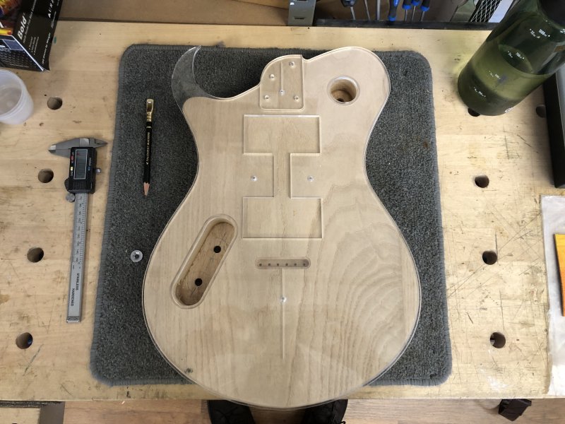 An unfinished guitar body on the workbench, over which sits a clear acrylic template. The outlines match, except in the top left where you can see the spikey horn shape has been remodelled to be a much more subtle shoulder.
