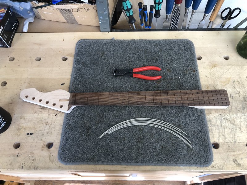 The neck on a workbench, next to some strips of fretwire and a cutting tool.