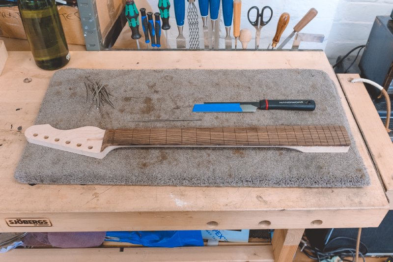 A work in progress guitar neck sits on the workbench: it has fret-slots cut in the fret board, but no frets. Besides the neck is a pile of short bits of fret wire, and a small hand saw.