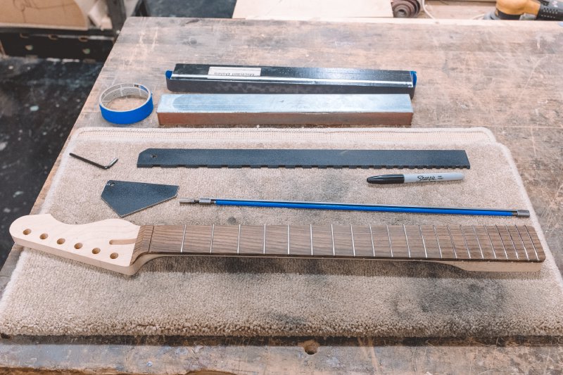 On the workbench we see the neck being worked on, next to which are: a truss-rod, a sharpie, an allen-key, a weird small metal plate with five sides of different lengths that is used for checking fret levels, a long notched ruler that is used to check the fretboard is level, two sanding bars with different grits of sandpaper on, and some masking tape.