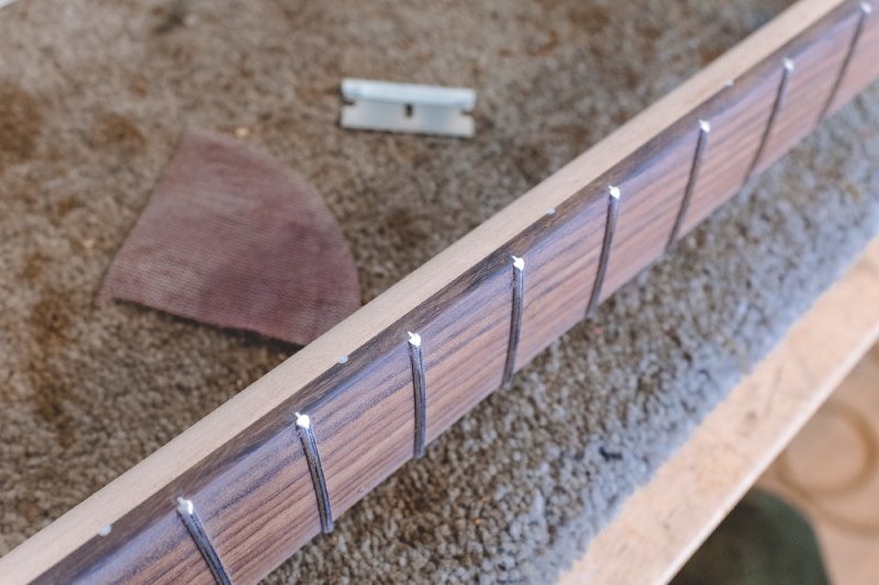 The neck is side up again, as we saw it when gluing earlier, but now the fret ends are flush with the side of the neck, and you can see the fretboard edge is slightly rounded too. Behind it sits a quater of a sanding disk and a razor blade.