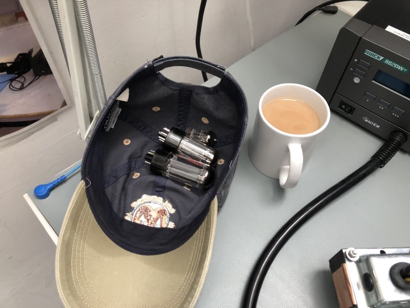 A baseball cap sits upturned on the workbench with three vacuum tubes/valves in it, and beside that is a fresh cup of tea.