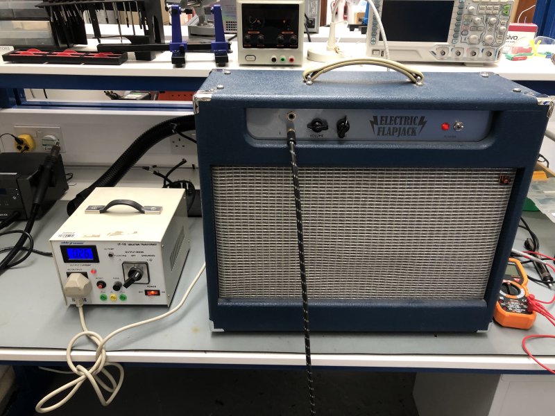 The amp is now back together, and seen front forward on the workbench, with a cable plugged into it. The amp is plugged not into the wall, but into a small grey box with switches and an LCD panel, which currently shows 0.26A being drawn. There is a red glow from the power indicator on the amp to show it is on.