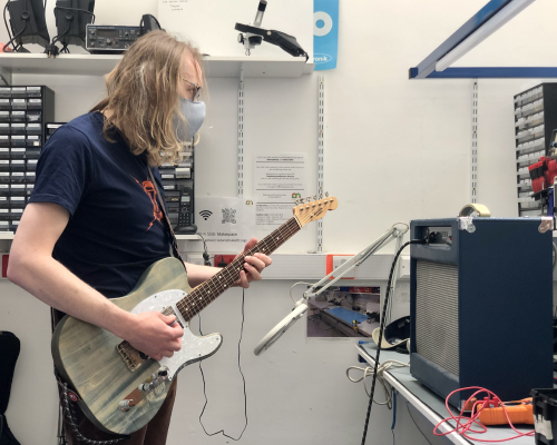 I'm stood next to the workbench with a blue guitar amp on it, and I have a guitar plugged into the amp for testing purposes. I'm staring at the amp as I play.
