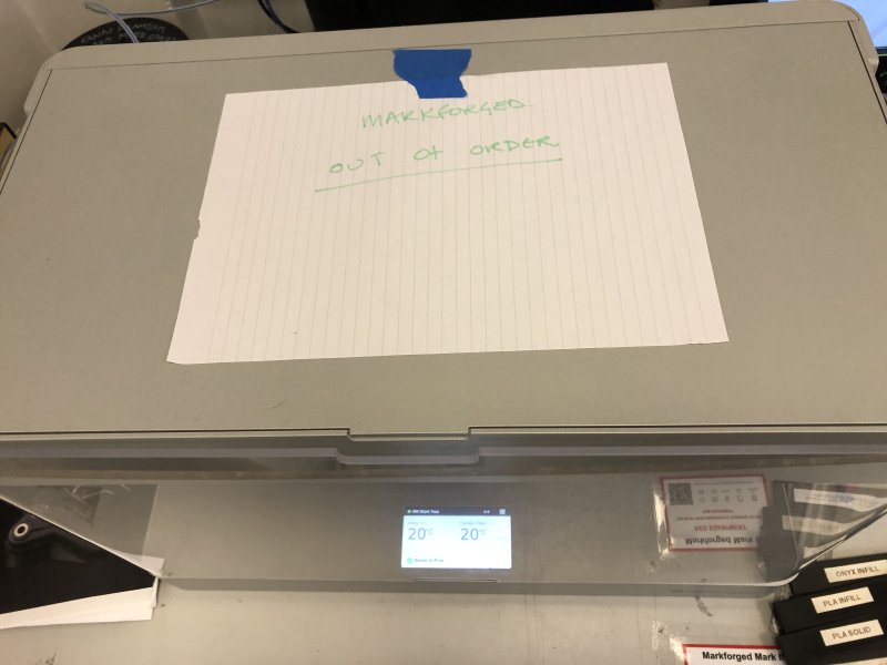 A boxy 3D-printer with a hand-written notice saying 'Markforged out of order' stuck to it.