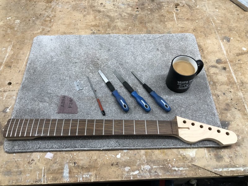 A guitar neck sits on a carpet-mat on a workbench, and beside it sit a set of small files, a razor blade, a sanding disk, and a cup of tea.