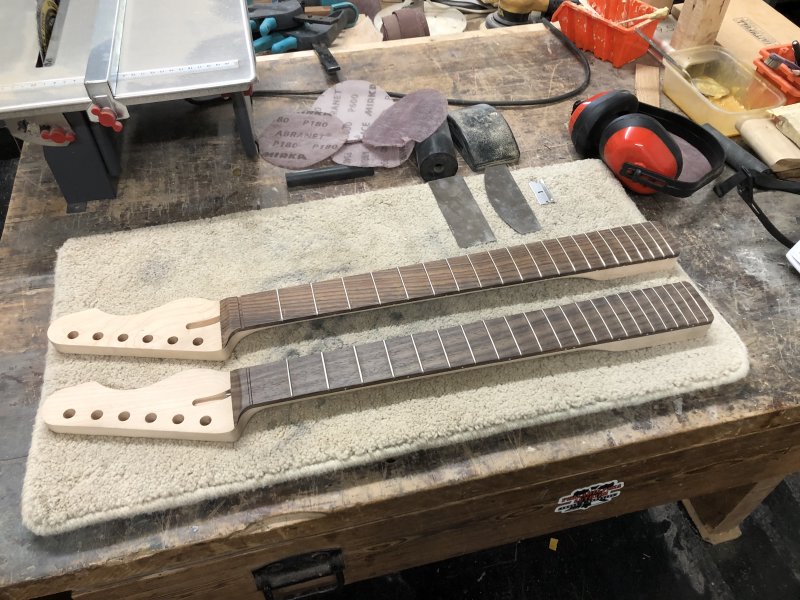 Two guitar necks sit on the workbench, both unfinished, but fretted and shaped. Both are maple necks with rosewood fretboards, but the colour of the rosewood is clearly different between the two, with one being more red. Close examination will show that one is slightly shorter than the other, and also has one additional fret.