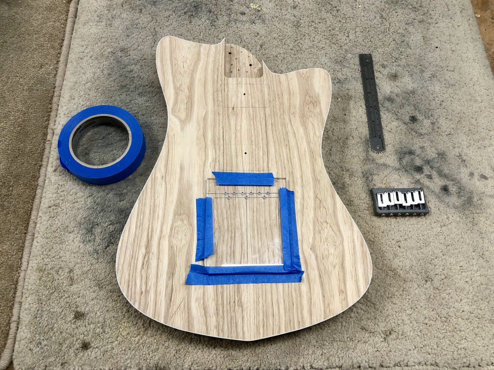 The shaped guitar body sits on the workbench, with a clear acrylic template taped to it with masking tape. The template has lines for the six string holes and three screw holes of the bridge. The bridge is seen off to the side.