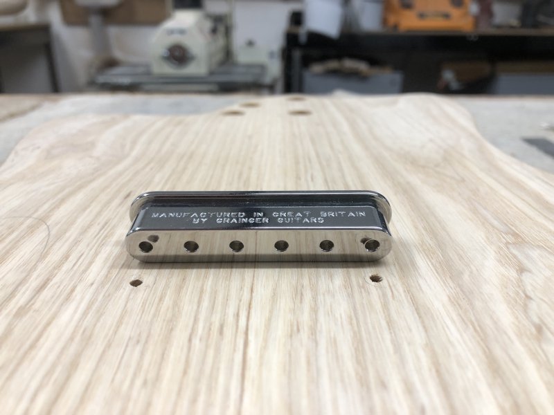 The rear of the guitar body on the workbench, showing the string-ferrule block, which is a oblong of metal with six holes in it, positioned next to the two holes that I drilled through to the back of the guitar.