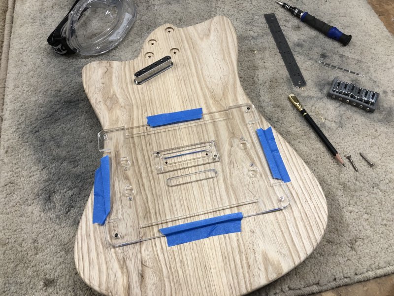 The guitar body sits on the a workbench, rear side up. Mounted to the back of it with masking table is a mess of clear acrylic in two layers, held together by metal pins.