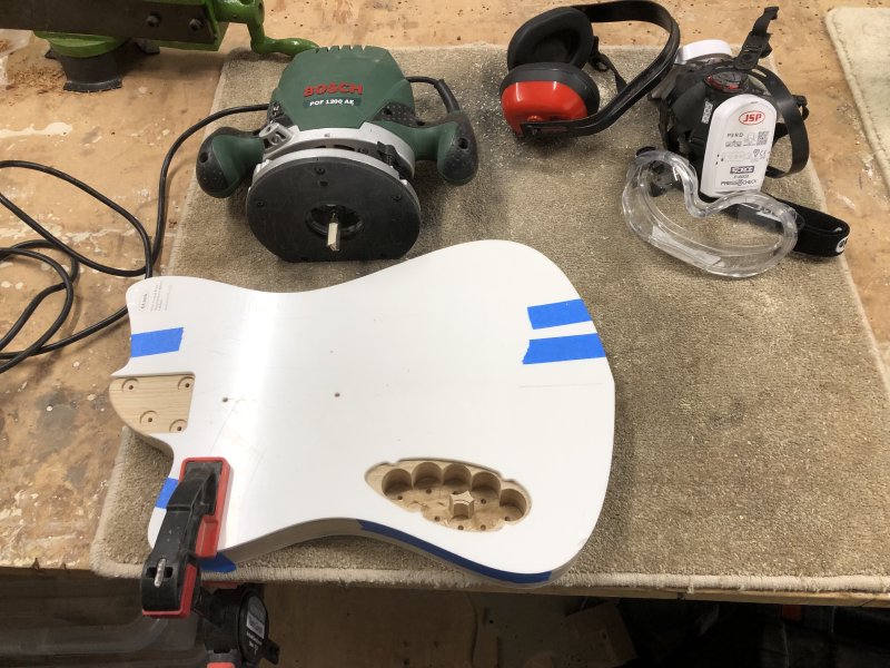 The guitar body is on the workbecnh with a white acrylic template on the back of it. There's one hole visible in the template (the other being under the clamp holding it all to the workbench), though which you can see one of the cluster of drilled holes. Next to it sits a plunge router and a pile of PPE.