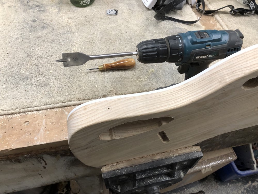 The guitar body is mounted in a vice, besides which is a power-drill with a large spade bit mounted in it.