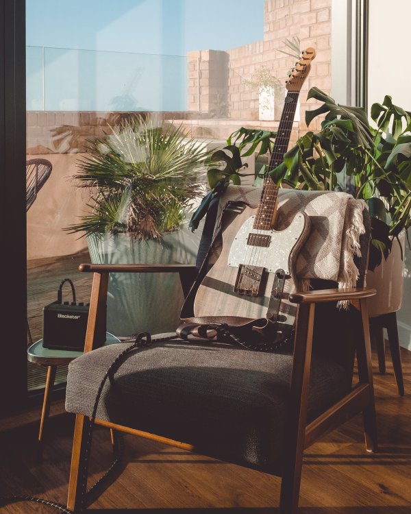 An electric guitar sits in a minimalist arm-chair, with some leafy plants behind it, and a small side stool to the left on which sits a tiny Blackstar portable practice amp.