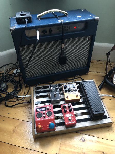 A blue coloured all-in-one amp sits in the corner of a room with a pedal board in front of it, and cables going everywhere behind it.